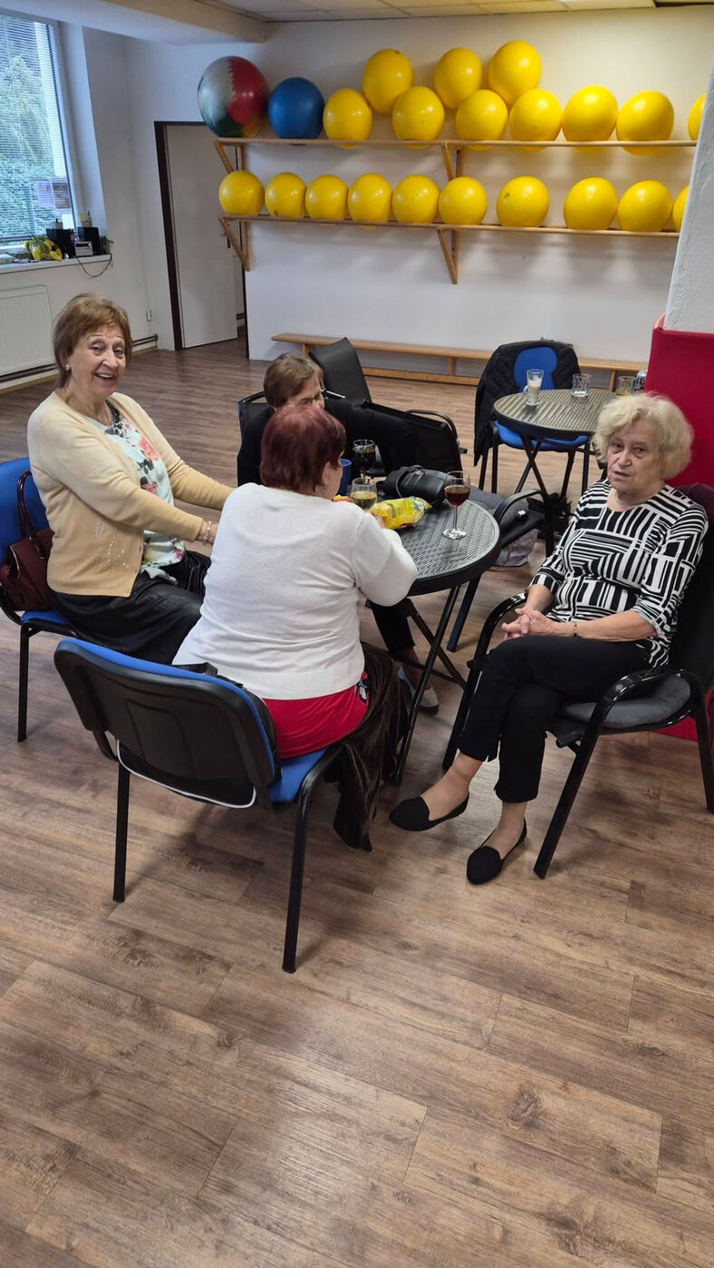 Čtyři ženy sedí u stolku a povídají si, za nimi police s gymnastickými míči