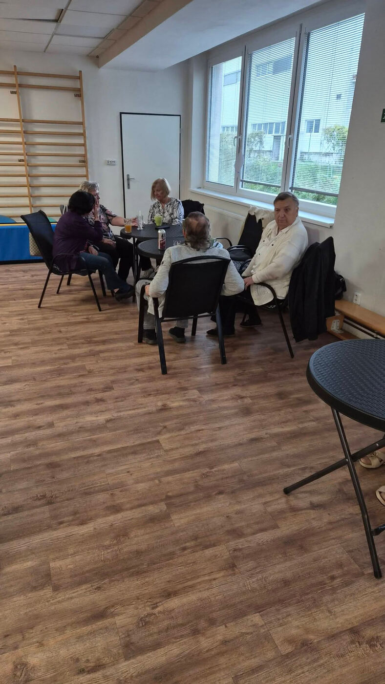 Skupinka seniorů u stolků u okna při společném posezení